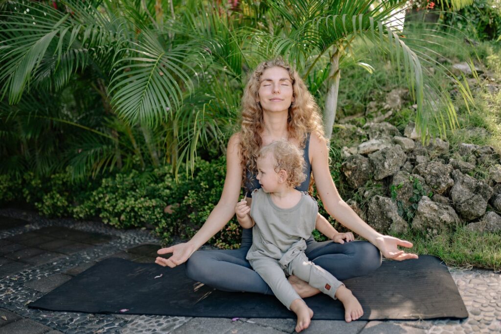 Full body happy relaxed mother sitting with eyes closed in lotus pose with cute boy on laps while practicing yoga in tropical green park