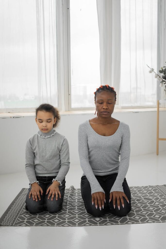 Full body of relaxed young African American woman with cute little daughter sitting on knees on floor and meditating together with closed eyes