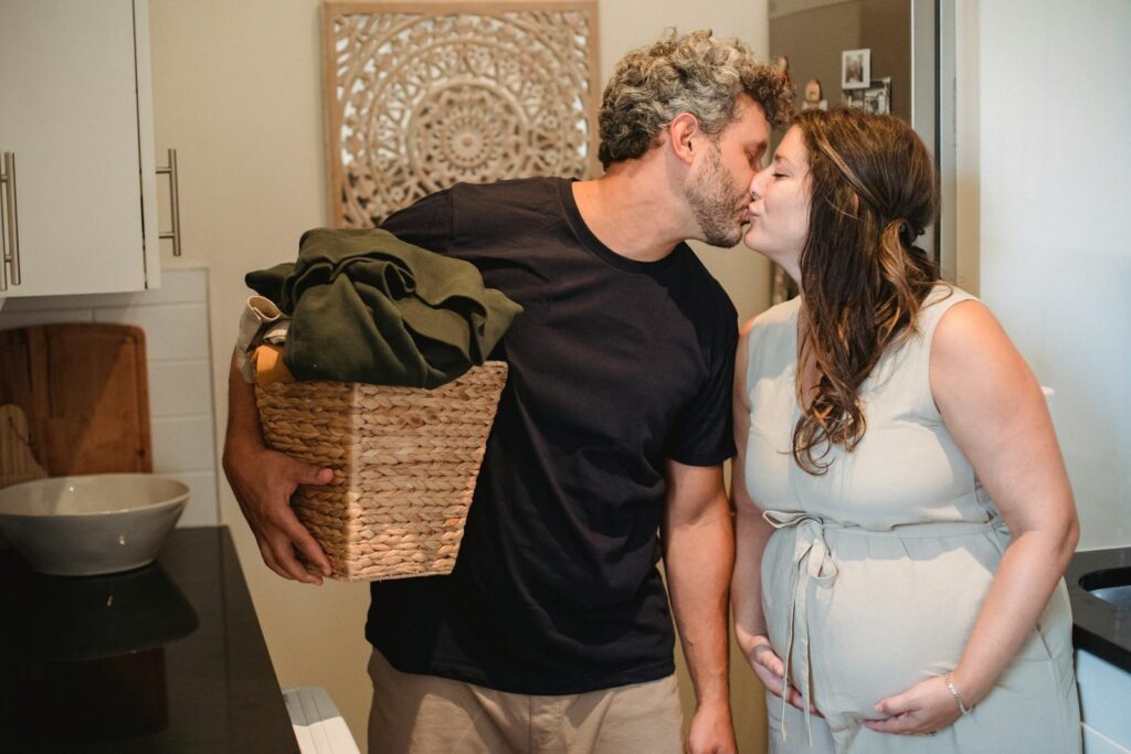 Happy adult man with laundry basket in hand kissing pregnant wife with closed eyes while standing together in bathroom