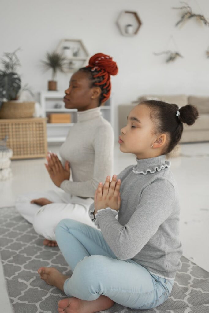 Side view of adorable little African American girl in casual clothes meditating with closed eyes with mother sitting on floor in Lotus pose